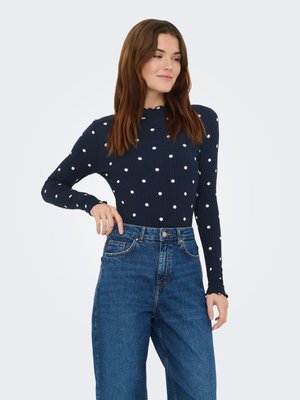 Marineblå langærmet top med hvide polkaprikker, ribdet tekstur og bølget halsudskæring, kombineret med højttaljede blå denimjeans.