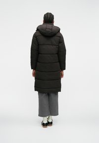 Melnas pūka jaka ar kapuci, ar quilted dizainu, garu garumu un izteiktu šuvju. Valkāta virs pelēkām platām biksēm un kedām.