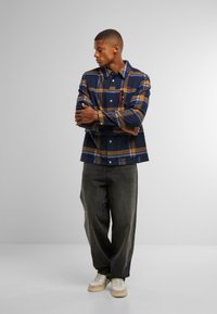 Marine rutete flanellskjorte med gule og blå striper, knapp foran og to brystlommer. Grå vide bukser og kremfargede joggesko.