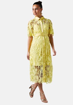 Donna che indossa un vestito midi in pizzo giallo con maniche corte, cintura e colletto, abbinato a sandali dorati con tacco alto e cinturini.