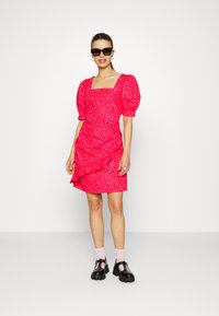 Vestido de mangas bufantes em rosa brilhante com padrão de rosas; decote quadrado e detalhes franzidos, combinado com sapatos de plataforma pretos e meias rosas.