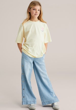 Geltona medvilninė marškinėliai su rausvu gėlių grafiniu vaizdu, dera prie mėlynų plačių džinsinių kelnių su šoninėmis sagomis ir atitrauktu kraštu.