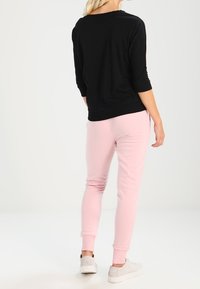 Schwarzes Oberteil mit drei Viertelärmeln, kombiniert mit hellrosa Jogginghosen. Die Jogginghosen haben umgekrempelte Knöchel, weichen Stoff und eine lockere Passform. Weiße Sneaker runden das Outfit ab.