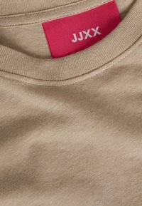 Hasselbruine katoenen t-shirt met een ronde hals. Voorzien van een rode label met witte tekst "JJXX." De stof heeft een gladde textuur en een subtiele glans.