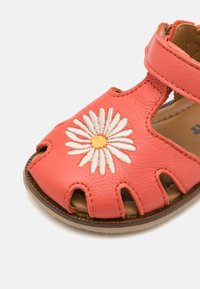 Korallfärgad lädersandal med utskärning och broderi av en vit prästkrage med en gul mitt. Kardborreband för säker fästning.