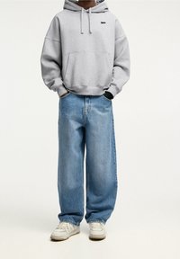 Harmaa hupullinen collegepaita, jossa etutasku ja logo; väljät, vaaleansiniset farkut; valkoiset lenkkarit, joissa vaaleanharmaat yksityiskohdat.