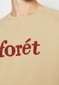 Sudadera de cuello redondo beige con letras rojas texturizadas que dicen "forét", llevada por una persona, recortada en el cuello y el pecho.