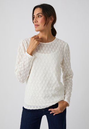 Mujer con cabello oscuro recogido en una coleta, que lleva una camiseta blanca de manga larga texturizada y pantalones azul oscuro, mirando hacia un lado con una mano cerca del cuello.