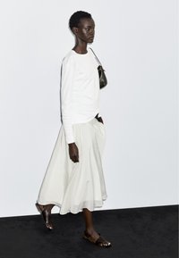 Hvid langærmet skjorte kombineret med et flydende hvidt skørt og brune sandaler; en lille mørk taske er hængt over skulderen.