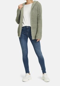 Tjockstickad cardigan i ljusgrön, kombinerad med en vit T-shirt och mörkblå smala jeans. Modellen har på sig vita sneakers.