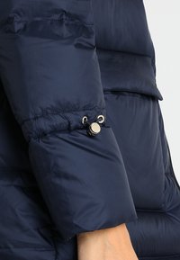 Rukáv námořnicky modré péřové bundy se stahovací manžetou, vybavený stříbrným patentním knoflíkem a hladkou lesklou texturou.