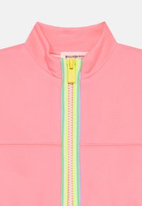 Ροζ νέον ζακέτα με φερμουάρ, που διαθέτει ψηλό γιακά, αντίθετο πράσινο φερμουάρ και ανοιχτά παστέλ στοιχεία κατά μήκος του φερμουάρ. Λεία υφή.