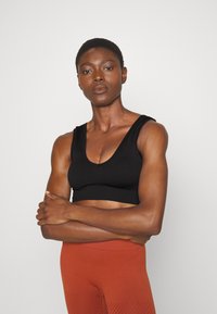 Top de desporto preto com nervuras, decote profundo em V e alças largas, utilizado com leggings na cor ferrugem com um padrão texturizado.