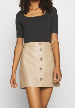 Vrouw met een zwart topje met korte mouwen en een beige leren minirok met knopen aan de voorkant, staand tegen een effen achtergrond.