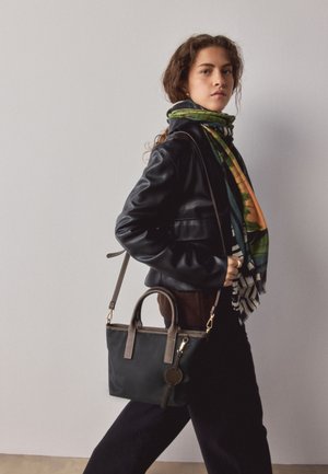 Žena kučeravými vlasmi, ktorá má na sebe čiernu koženú bundu a farebný šál, nesie čiernu kabelku s hnedými popruhmi cez rameno.