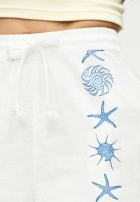 Λευκά σορτς με κορδόνι και μπλε σχέδια με θέμα τη θάλασσα, που περιλαμβάνουν αστερία, κοχύλι και έναν αγκαθωτό αχινό κατά μήκος της πλευράς.