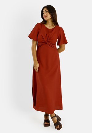Femme debout portant une robe maxi couleur rouille avec des manches courtes volantées et un détail twist à l'avant, associée à des sandales compensées marron foncé.
