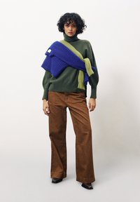 Groene gebreide trui met hoge nek, gecombineerd met bruine wijde corduroy broeken. Blauwe en groene gebreide omslagdoek over de schouders.