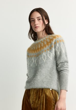 Junge Frau mit langen braunen Haaren trägt einen grauen Pullover mit orange-weißen Mustern und braune Samthose, steht vor einem schlichten Hintergrund.