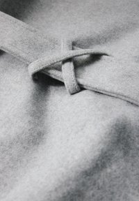 Szary, filcowy materiał o teksturowanej powierzchni, z detalem w postaci węzła na pasku i gładkimi krawędziami, tworzący miękki, warstwowy wygląd.