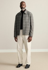 Gray plaid jacket with a collared neckline, worn over a black turtleneck. Paired with cream-colored trousers and black shoes.