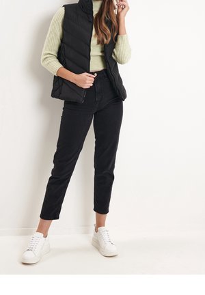 Femme portant une doudoune noire, un pull côtelé vert clair, un jean noir taille haute et des baskets blanches, debout contre un mur blanc.