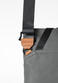 Gray fabric bag detail with a black strap and brown leather buckle. Features a textured surface and minimal stitching. Adjustable strap length.