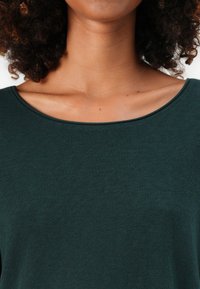Woman with curly hair wearing a dark green scoop-neck top, showing neck and collarbone area against a plain background.