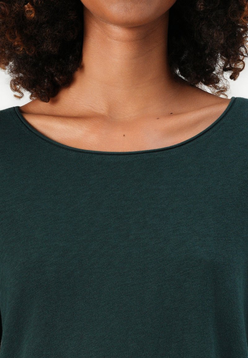 Frau mit lockigem Haar, die ein dunkelgrünes Top mit Rundhalsausschnitt trägt, das den Hals- und Schlüsselbeinbereich zeigt, vor einem einfarbigen Hintergrund.