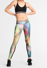 Kobieta w czarnym biustonoszu sportowym i legginsach w wielokolorowy abstrakcyjny wzór, stojąca z rękami na biodrach na tle białym.