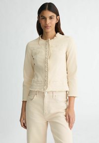 Giacca di denim beige con bordi sfrangiati, maniche lunghe e due tasche sul petto. Presenta una chiusura anteriore intrecciata e un design aderente.