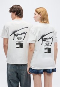 Δύο άτομα με την πλάτη, φορούν λευκά T-shirt Tommy Jeans με μαύρα ονόματα πόλεων και τυπώματα λογότυπου στην πλάτη.