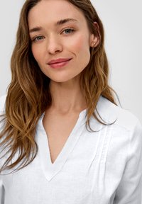 Blouse en lin blanc avec un décolleté en V et des accents plissés. La texture est douce et légère, adaptée à un port décontracté. Design minimaliste.