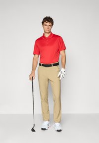 Muž v červenom polo tričku, béžových nohaviciach, bielom golfovom rukavici a bielych topánkach, ktorý drží golfovú palicu na neutrálnom šedom pozadí.
