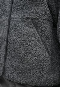 Γκρι φλις μπουφάν με υφή, διαθέτει πλαϊνή τσέπη και μαύρο περίγραμμα κατά μήκος των άκρων για αντίθεση και ανθεκτικότητα.