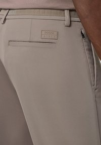 Pantaloni beige con una texture liscia, dotati di una tasca posteriore e di una piccola etichetta con il logo "BOGGI BTECH". Dettaglio con tasca laterale con zip.