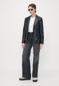 Černý blazer z umělé kůže přes bílé tričko, spárovaný s tmavými džínami s vysokým pasem a zelenými platformovými botami, se stylovými oválnými slunečními brýlemi.