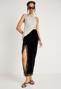 Aszimmetrikus bézs felső és fekete bársonyszoknya csipkebetéttel, oldalsó hasítékkal. A modellen fekete napszemüveg és pántos fekete szandál van.