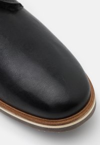 Zapato de cuero negro con textura suave, puntera redondeada y suela contrastante en marrón y blanco. Diseño minimalista sin herrajes visibles.