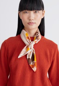Mujer con cabello negro liso lleva un suéter naranja y una bufanda de seda con estampado floral atada al cuello contra un fondo sencillo.