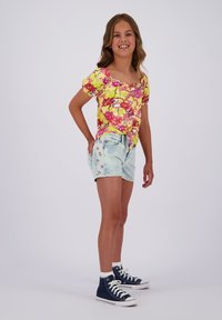 Helles, gelbes Blumenoberteil mit kurzem Arm, kombiniert mit hellen Denim-Shorts, die mit gestickten Blumen verziert sind und einen lässigen Schnitt haben. Abgerundet mit marineblauen Sneakers.