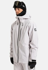 Mann in einer hellgrauen wasserdichten Jacke, mit schwarzen Handschuhen, schwarzer Mütze und Skibrille, die nach links zeigt, vor einem weißen Hintergrund.