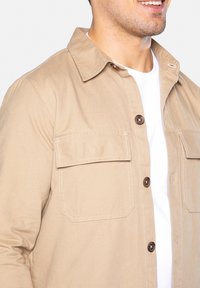 Beige skjortejakke lavet af letvægtsstof, med to forlommer, brune knapper og kravedesign.
