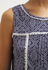 Mörkblå utan ärmar topp med vit spets och pom-pom kant. Har intrikata vita geometriska och blommiga mönster i hela tyget.