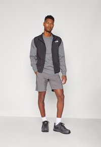 Zwarte vestjasje over een grijze sweatshirt, grijze shorts, witte sokken en grijze wandelschoenen. Opvallend merkje op het jasje.