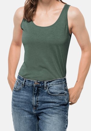Vrouw die een groen mouwloos hemdje draagt, ingestopt in een high-waisted blauwe spijkerbroek met handen in de zakken.