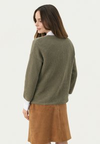 Oliwkowy sweter z dzianiny o prążkowanej teksturze, luźny krój i opadające ramiona, zestawiony z brązową spódnicą z zamszu. Widoczny biały kołnierzyk koszuli.