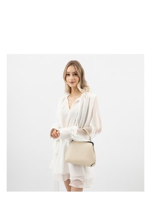 Jonge vrouw in witte gelaagde jurk met een beige handtas tegen een effen witte achtergrond, zacht glimlachend.