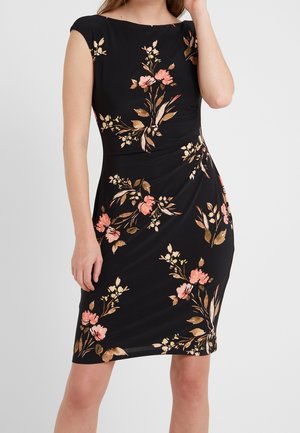 Mujer con vestido negro sin mangas hasta la rodilla con estampados florales en rosa y marrón, posando con la mano cerca del rostro.