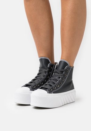 Jambes portant des baskets montantes noires à plateforme avec des semelles épaisses blanches texturées et des lacets noirs sur fond blanc.